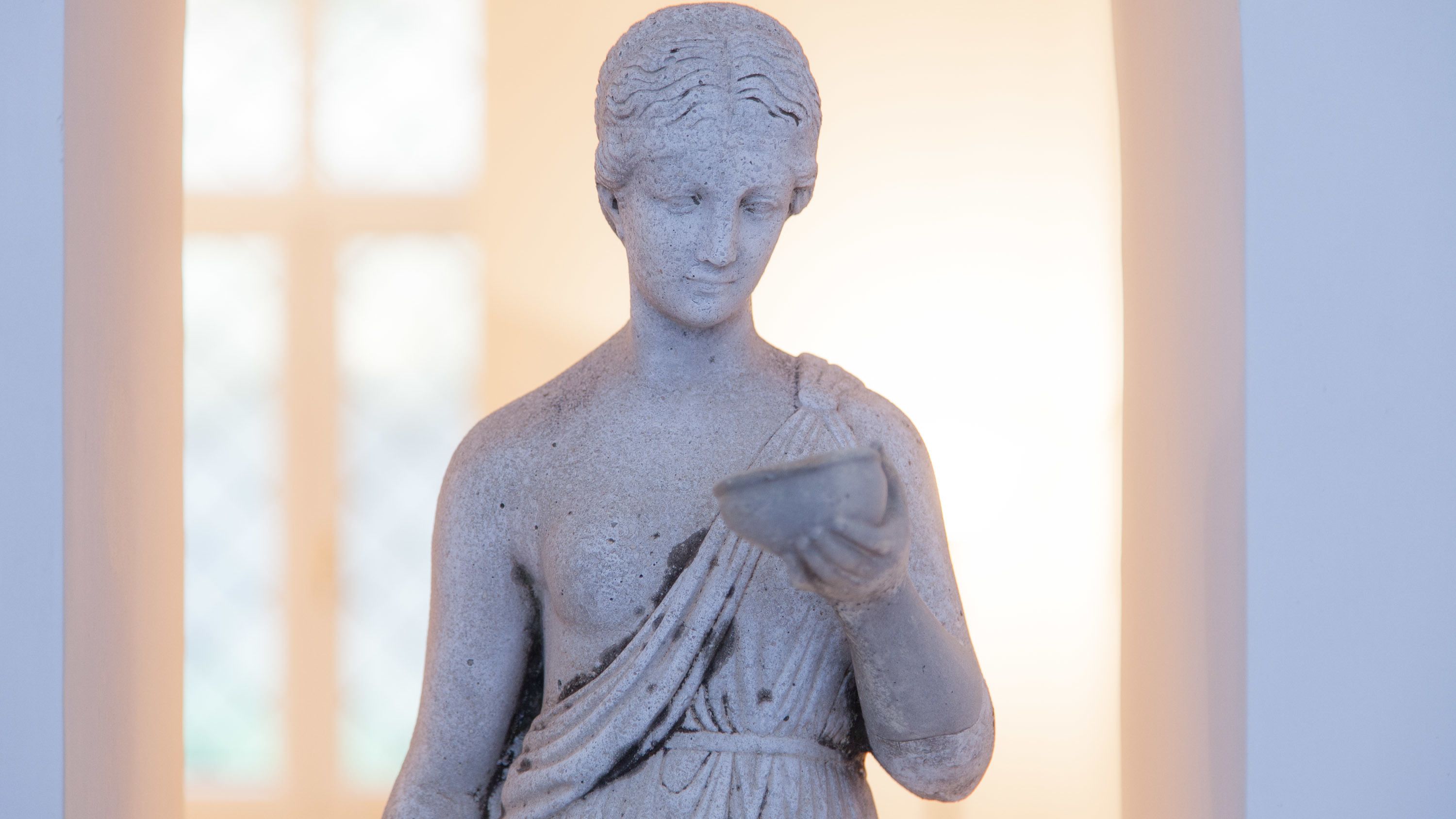 Eine Marmorstatue einer Figur, die ein kleines Objekt hält, vor einem sanft beleuchteten Hintergrund mit einem Fenster.