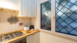 Eine Küche mit Gasherd, Wasserkocher und einem Teller mit einem Croissant. Auf der Arbeitsfläche stehen zwei Tassen, und ein Fenster mit Rautenmuster ist zu sehen.