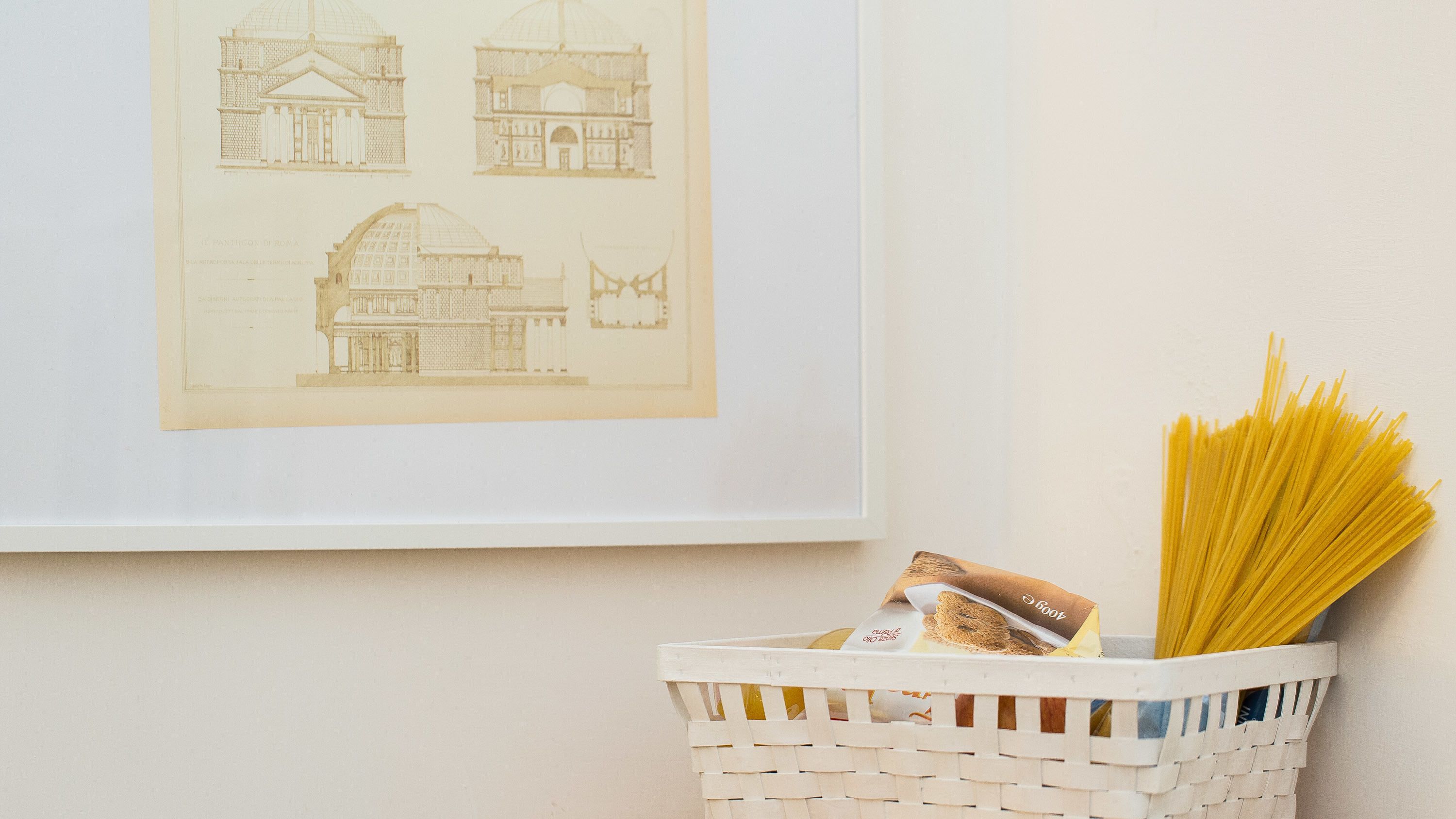 A framed architectural drawing on a wall, a basket with pasta and a package, and a white wall.