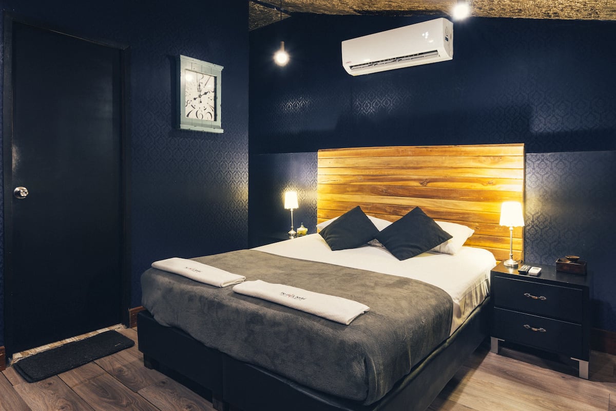 A modern hotel room with a large bed, dark walls, a wooden headboard, a bedside table with a lamp, and a small nightstand.