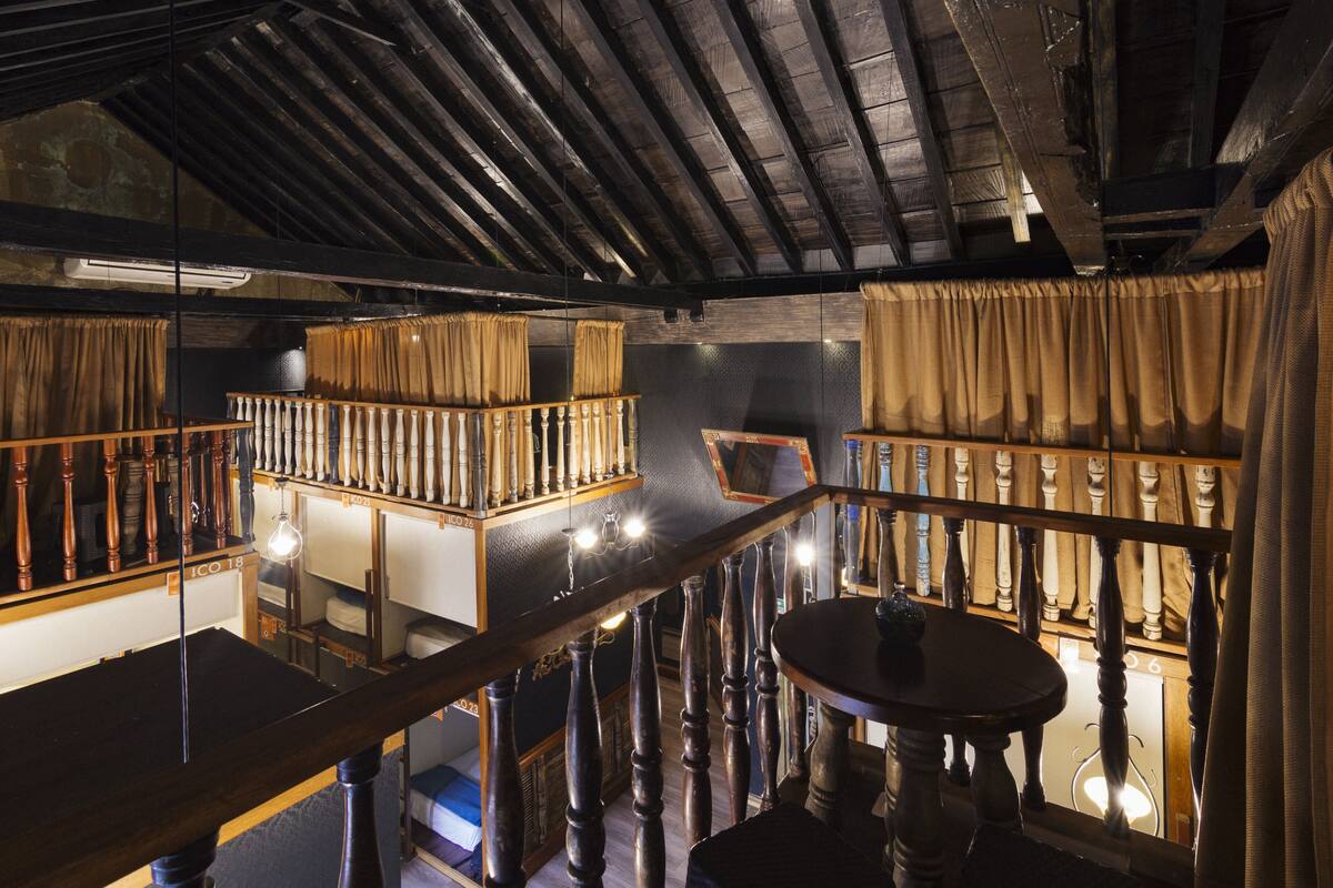 A room with a wooden staircase, a round table, and a view of a balcony with curtains.