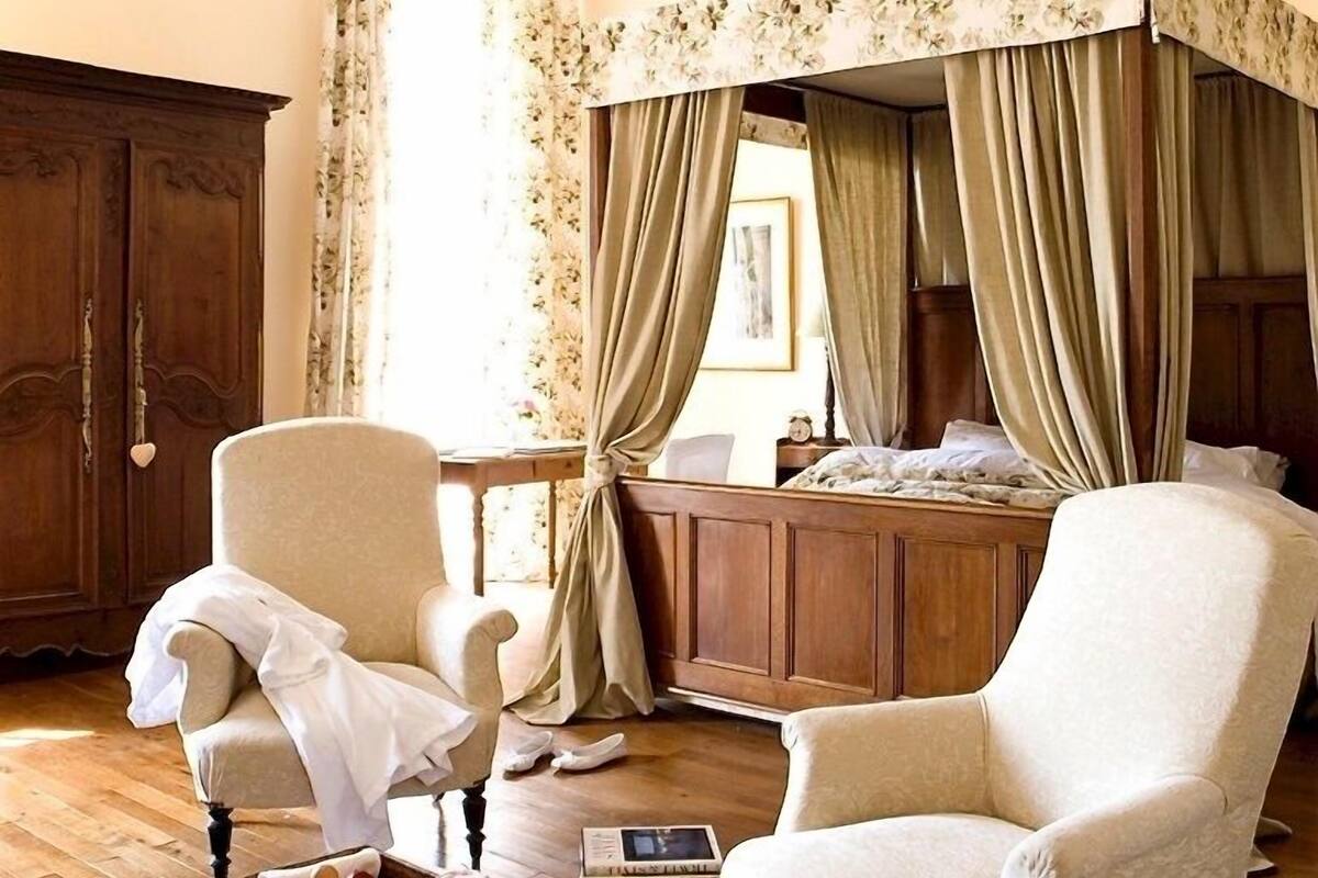 Una habitación con un armario de madera, dos sillones, una mesita de noche con un libro y una cama con cortinas.
