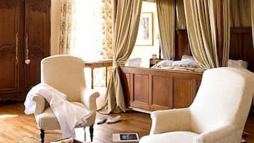 A room with a wooden wardrobe, two armchairs, a bedside table with a book, and a bed with curtains.