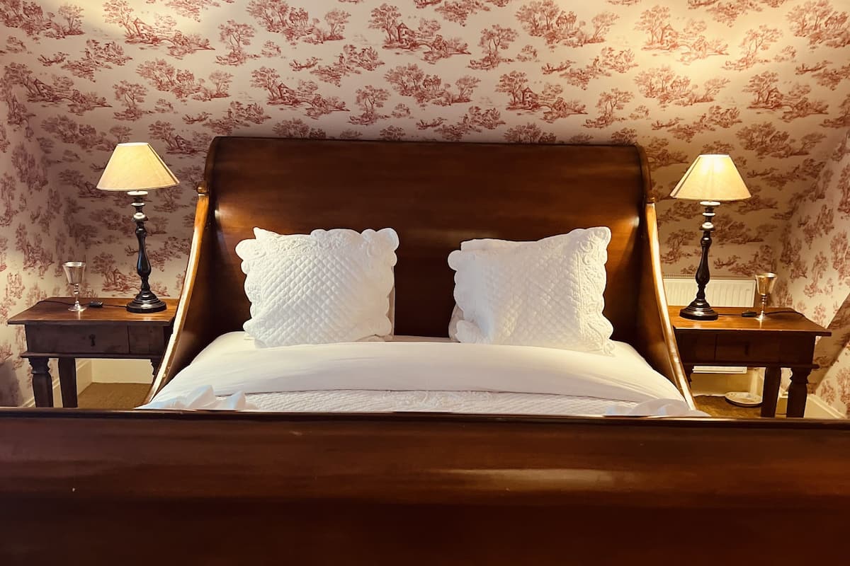 Una cama bien tendida con sábanas blancas, flanqueada por dos mesitas de noche con lámparas, en una habitación con papel tapiz floral.