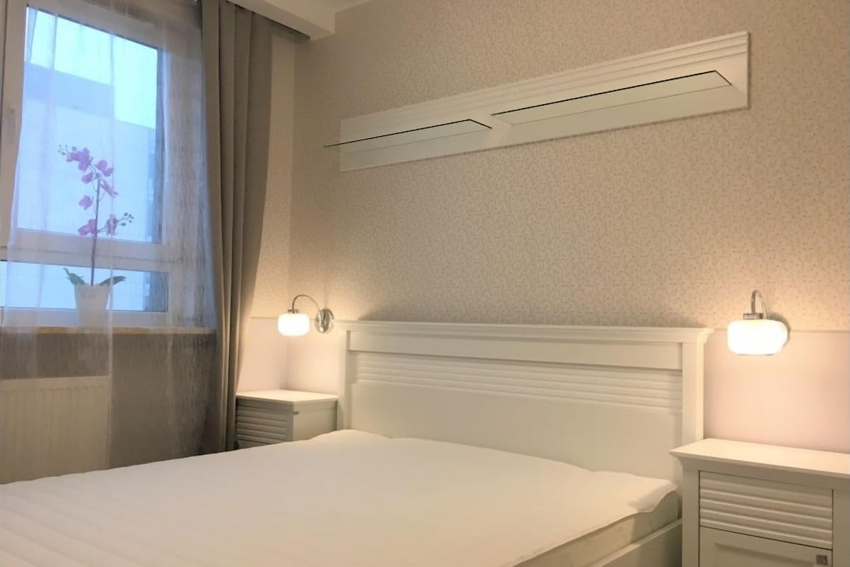 A bedroom with a bed, two bedside tables, a wall-mounted air conditioner, and a window with curtains.