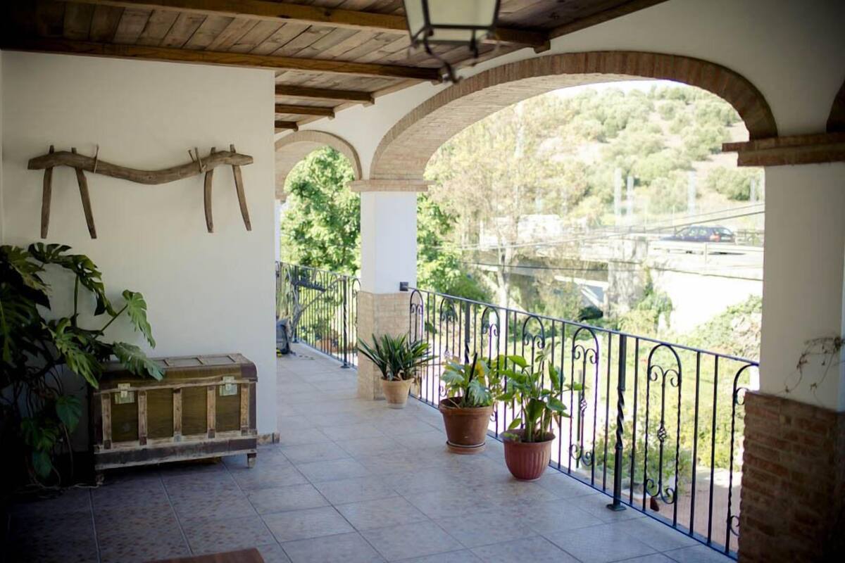 A covered outdoor area with a wooden bench, potted plants, and a view of a bridge and trees.