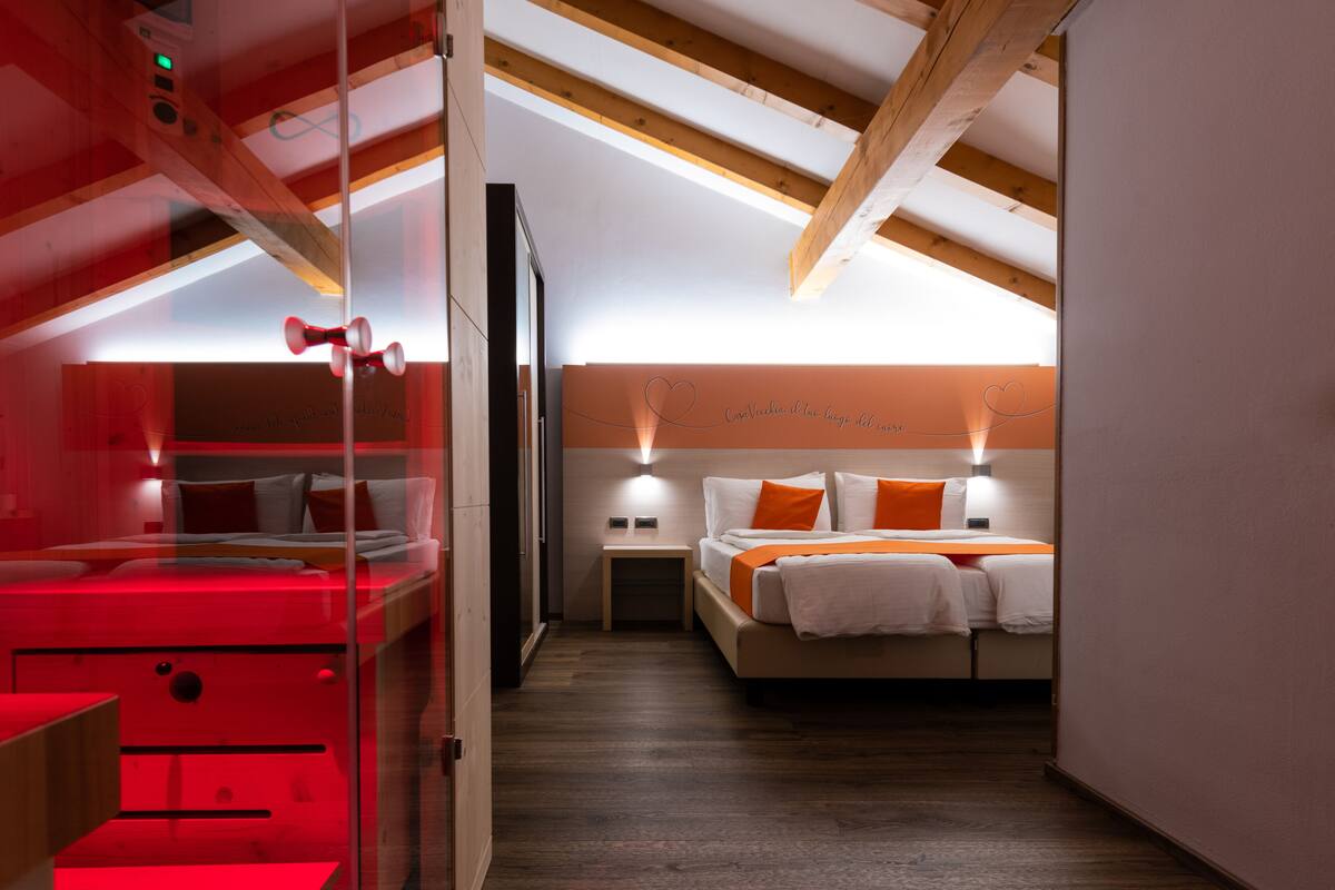 A modern hotel room with a glass partition, wooden beams, a bed with white and orange bedding, and a wall-mounted light fixture.