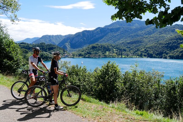 Two cyclists on a paved path with a lake and mountains in the background.