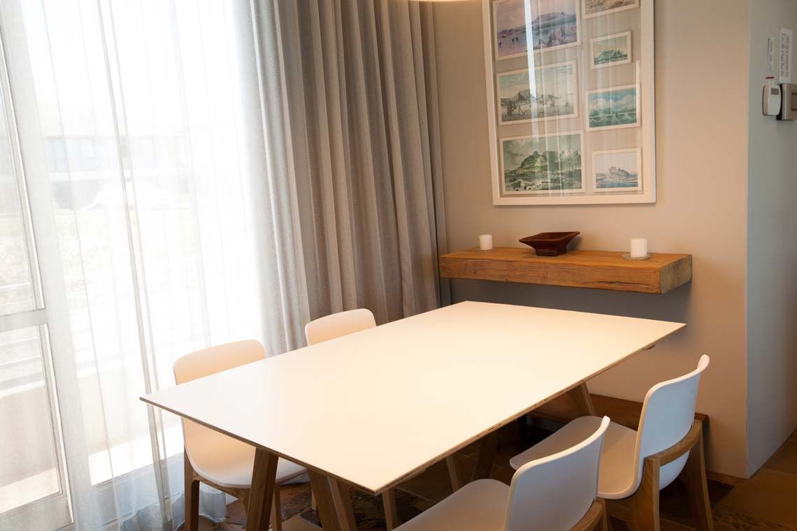 A modern dining area with a white table and chairs, a wooden shelf with decorative items, and a window with curtains.