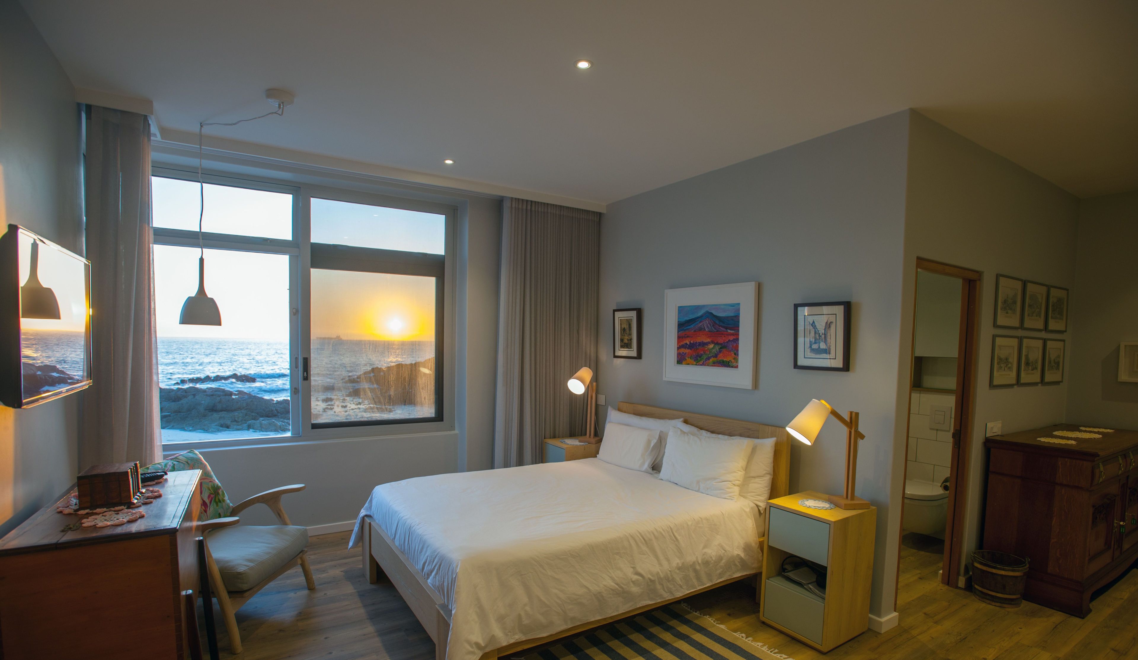 A hotel room with a large window overlooking the sea, a bed, a desk with a chair, a bedside table with a lamp, and artwork on the walls.