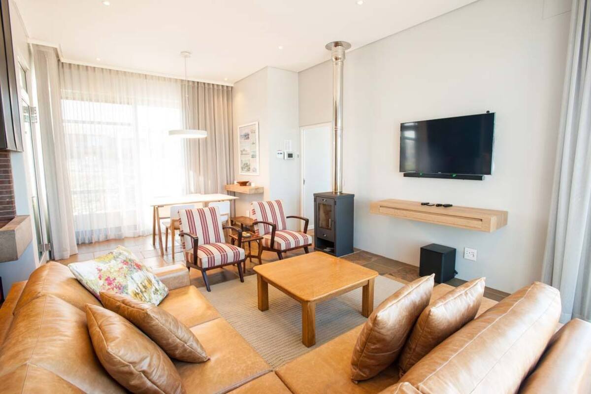 Un salon moderne avec un canapé en cuir, une table basse en bois, des fauteuils à rayures et un téléviseur à écran plat fixé au mur.