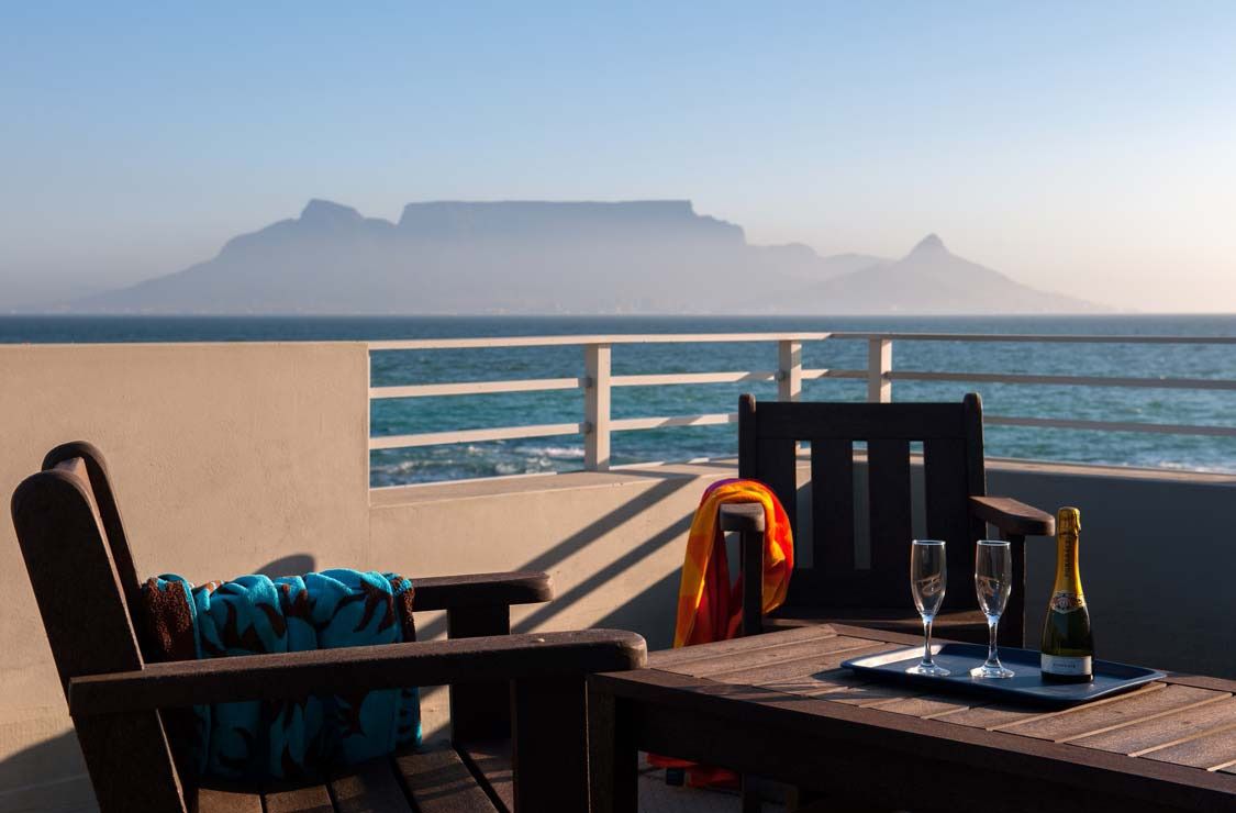 A wooden table with a bottle of champagne and two glasses on a tray, overlooking a coastal view with a chair and a colorful towel.