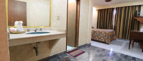 A hotel room with a bathroom featuring a sink, mirror, and towel rack. The room has a bed with a colorful bedspread, a desk, and a chair.