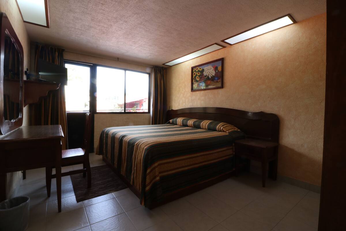 A bedroom with a bed, a desk, a chair, a window, and a painting on the wall.