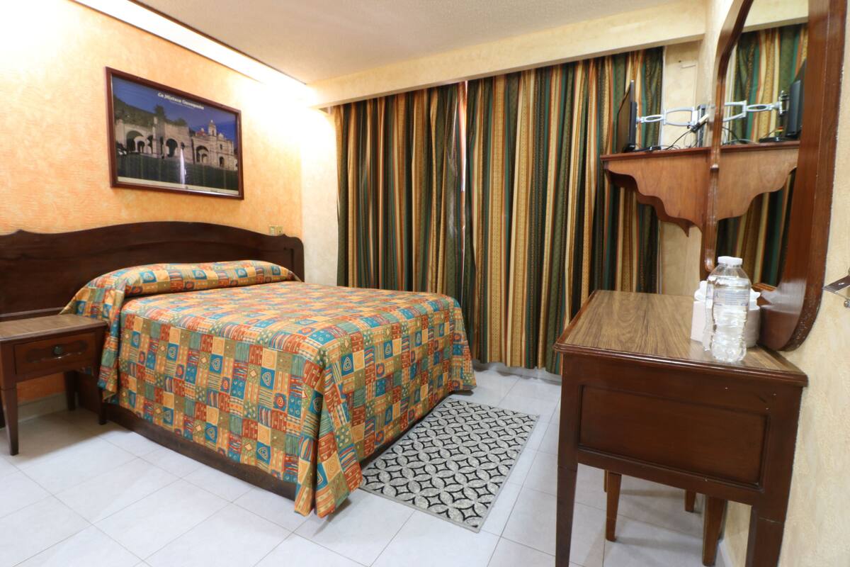 A hotel room with a bed, a bedside table, a desk with a mirror, and a patterned rug.