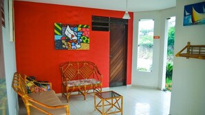 Una habitación con una pared roja, una puerta de madera y una ventana. Hay dos sillas de mimbre y una mesita.