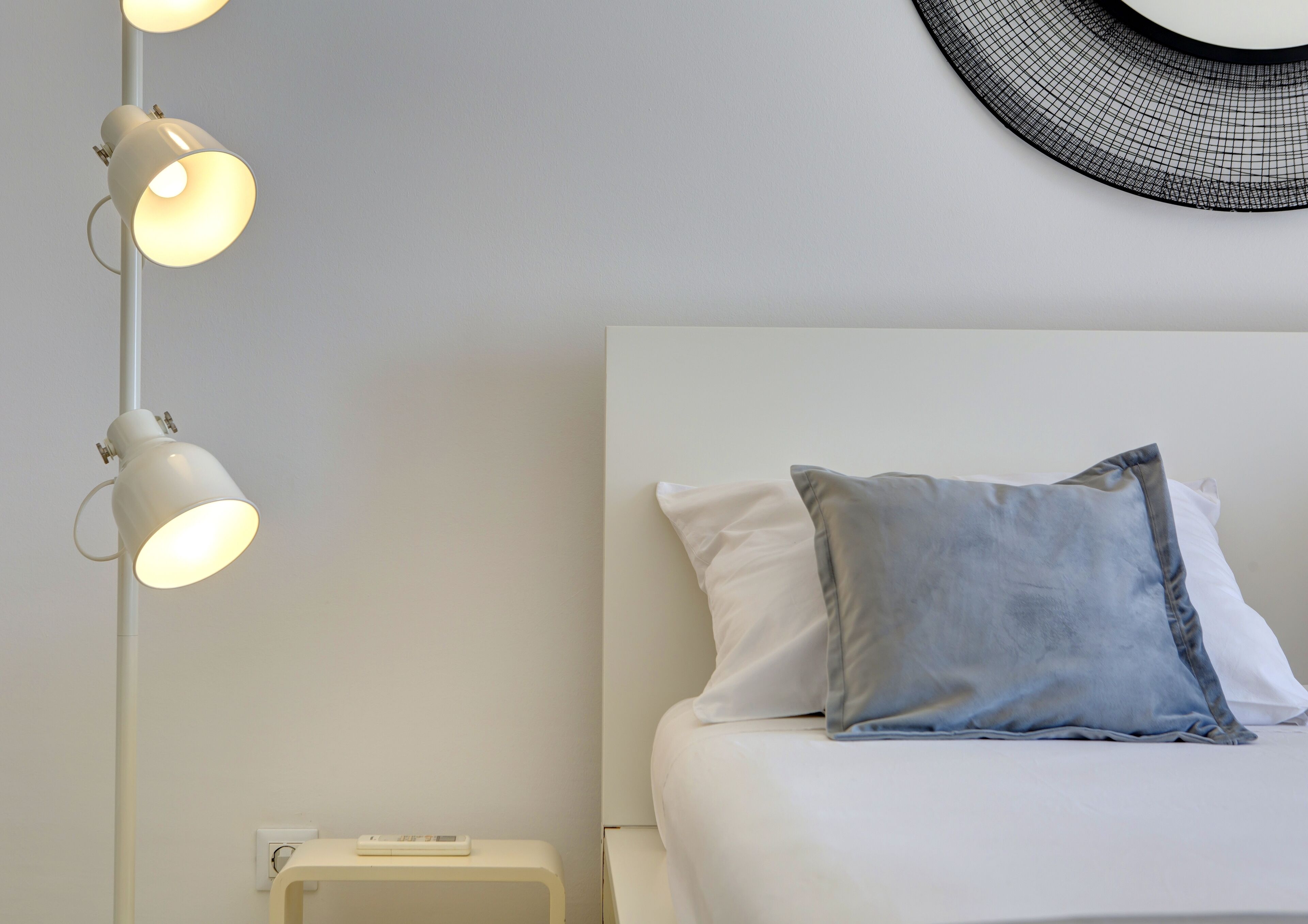 Una cama bien hecha con almohadas blancas y grises, una mesita de noche blanca con un pequeño dispositivo electrónico, y una lámpara de pie moderna con varias bombillas.