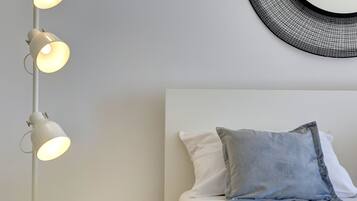 Una cama bien hecha con almohadas blancas y grises, una mesita de noche blanca con un pequeño dispositivo electrónico, y una lámpara de pie moderna con varias bombillas.