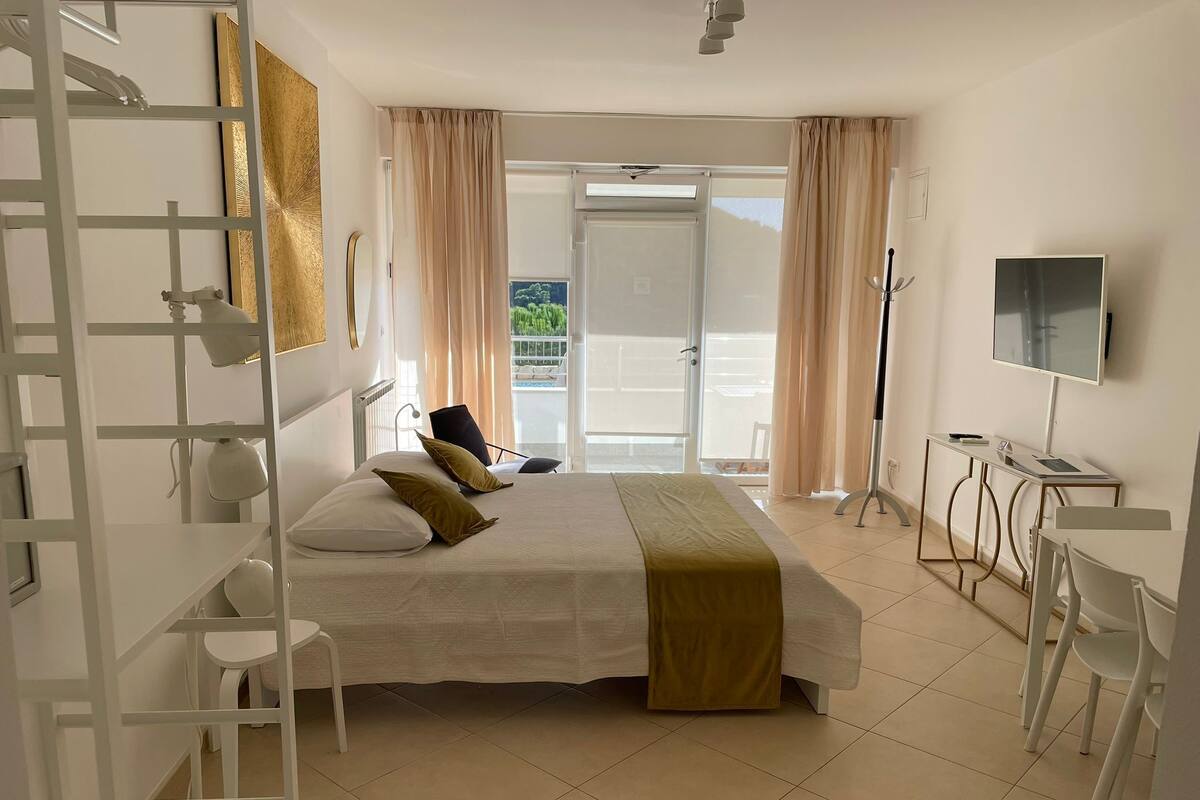 A hotel room with a bed, a desk, a chair, a TV, and a balcony with curtains.