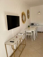 A modern, minimalist room with a flat-screen TV mounted on the wall, a small table with a remote control, a white dining table with chairs, a microwave, and a toaster on the counter.