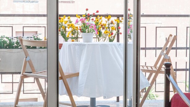 A table with a white tablecloth, a potted plant with yellow and pink flowers, and wooden chairs. - Rental In Rome Guinizzelli (Rome)