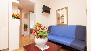 A living room with a blue sofa, a white vase with flowers, a small table, a television mounted on the wall, and a door leading to another room. - Rental In Rome Guinizzelli (Rome)