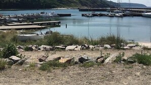 Ein Yachthafen mit vertäuten Booten, eine Mole, die in ruhiges Wasser ragt, und eine hügelige Landschaft im Hintergrund.