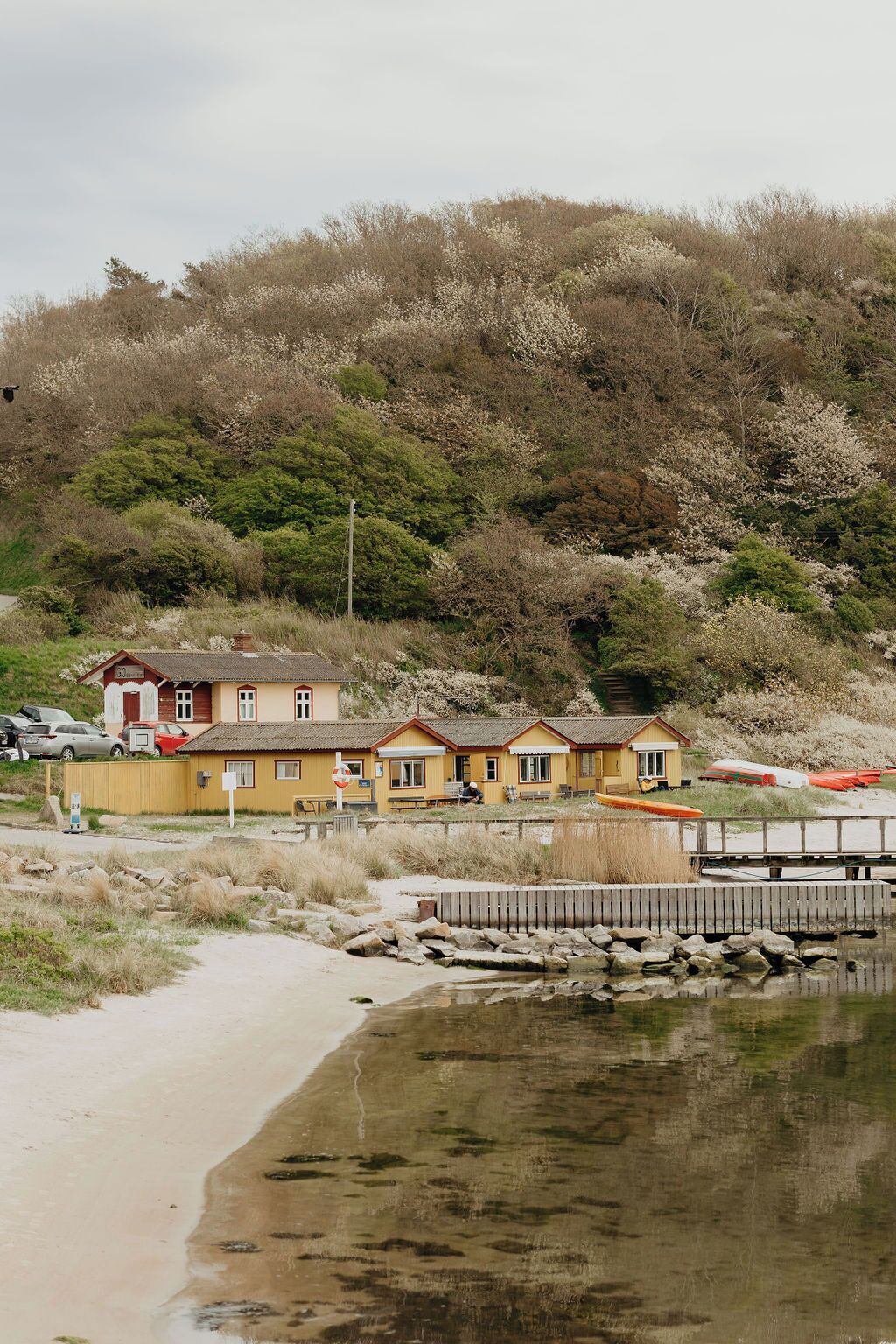 A row of yellow buildings with a beach in the foreground and a hill with trees in the background.