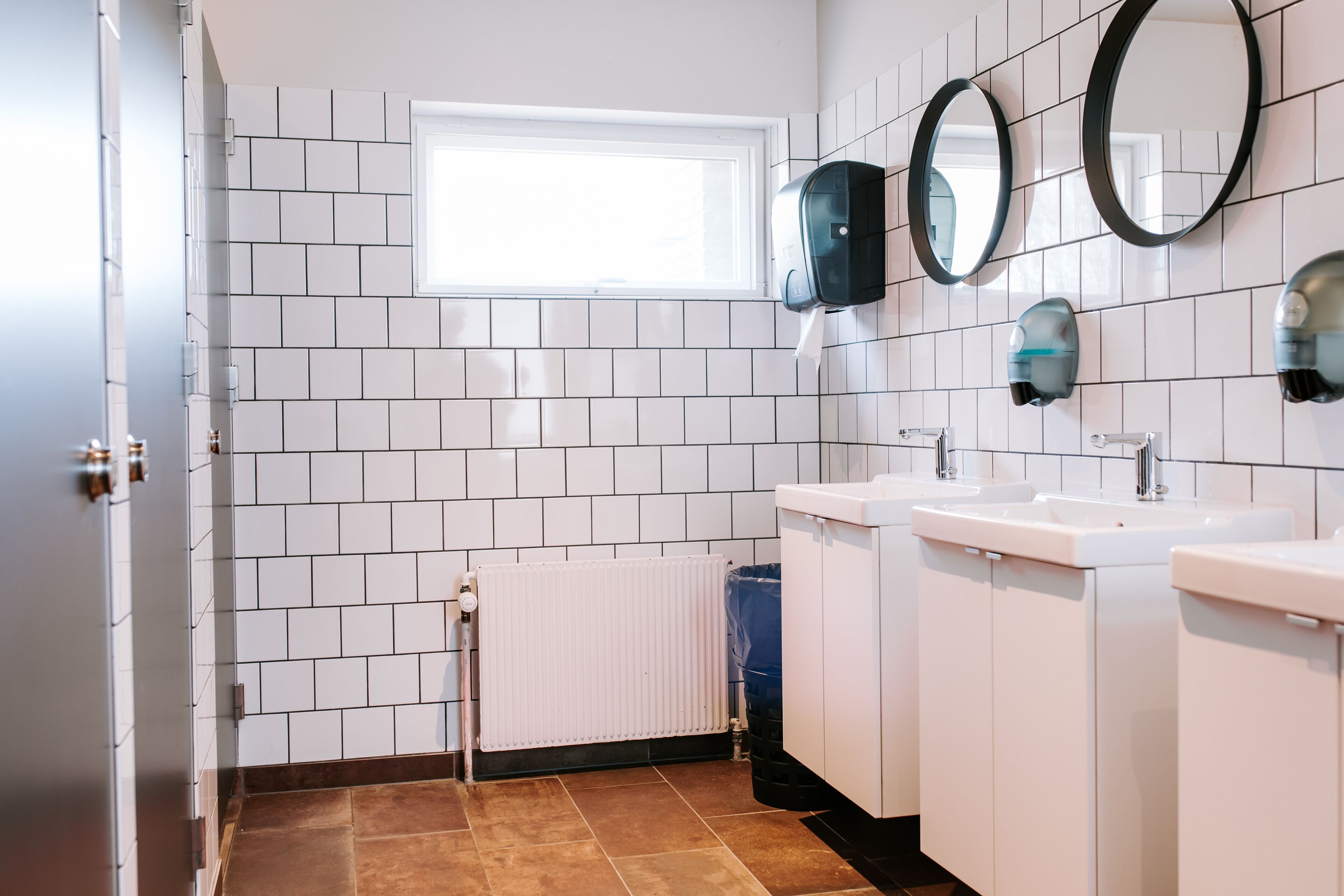 Um banheiro moderno com paredes revestidas de azulejos brancos, uma janela, três pias e duas espelhas fixadas na parede.