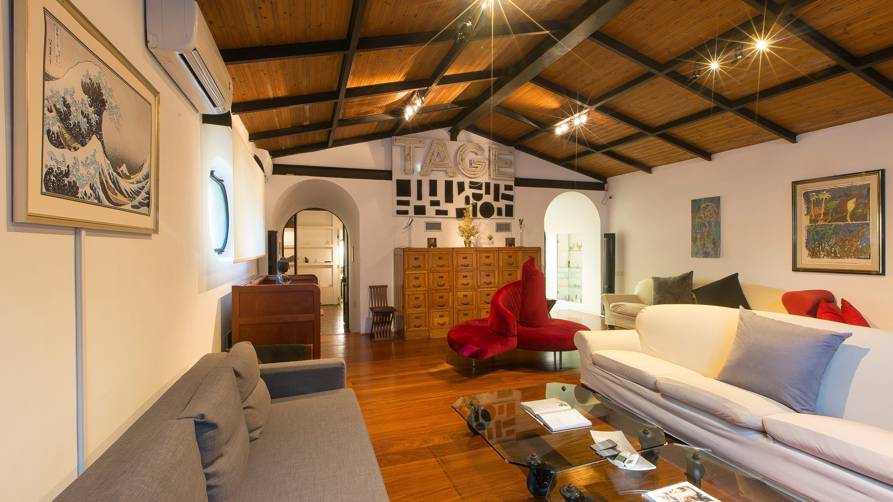 Uma sala de estar ampla com teto alto, vigas de madeira, um sofá, uma poltrona vermelha e uma mesa de centro de vidro.