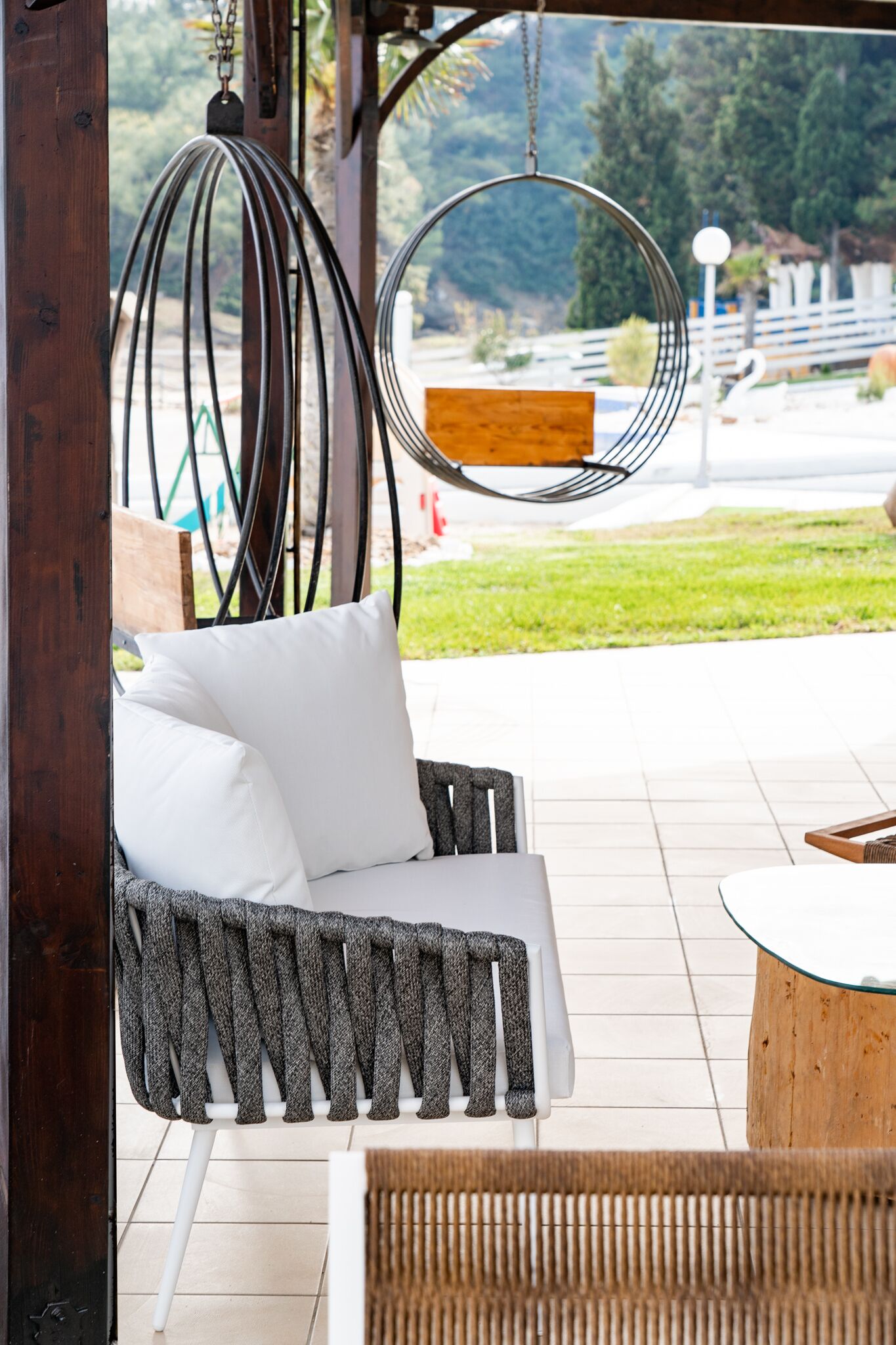 Une chaise en osier avec des coussins blancs et une balançoire suspendue en métal, toutes deux placées sur une terrasse carrelée offrant une vue sur une pelouse et des arbres.
