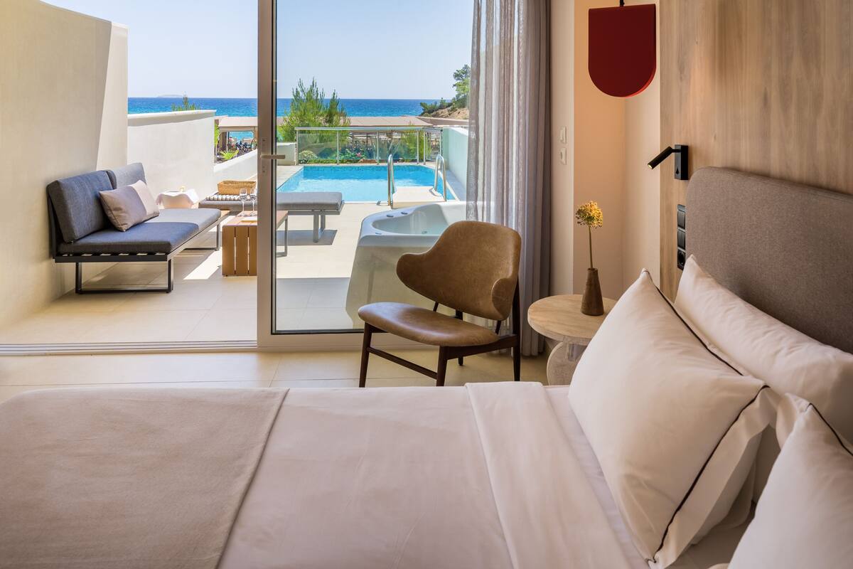 Quarto de hotel com uma cama grande, um sofá, uma poltrona, uma mesinha e vista para a piscina e praia.
