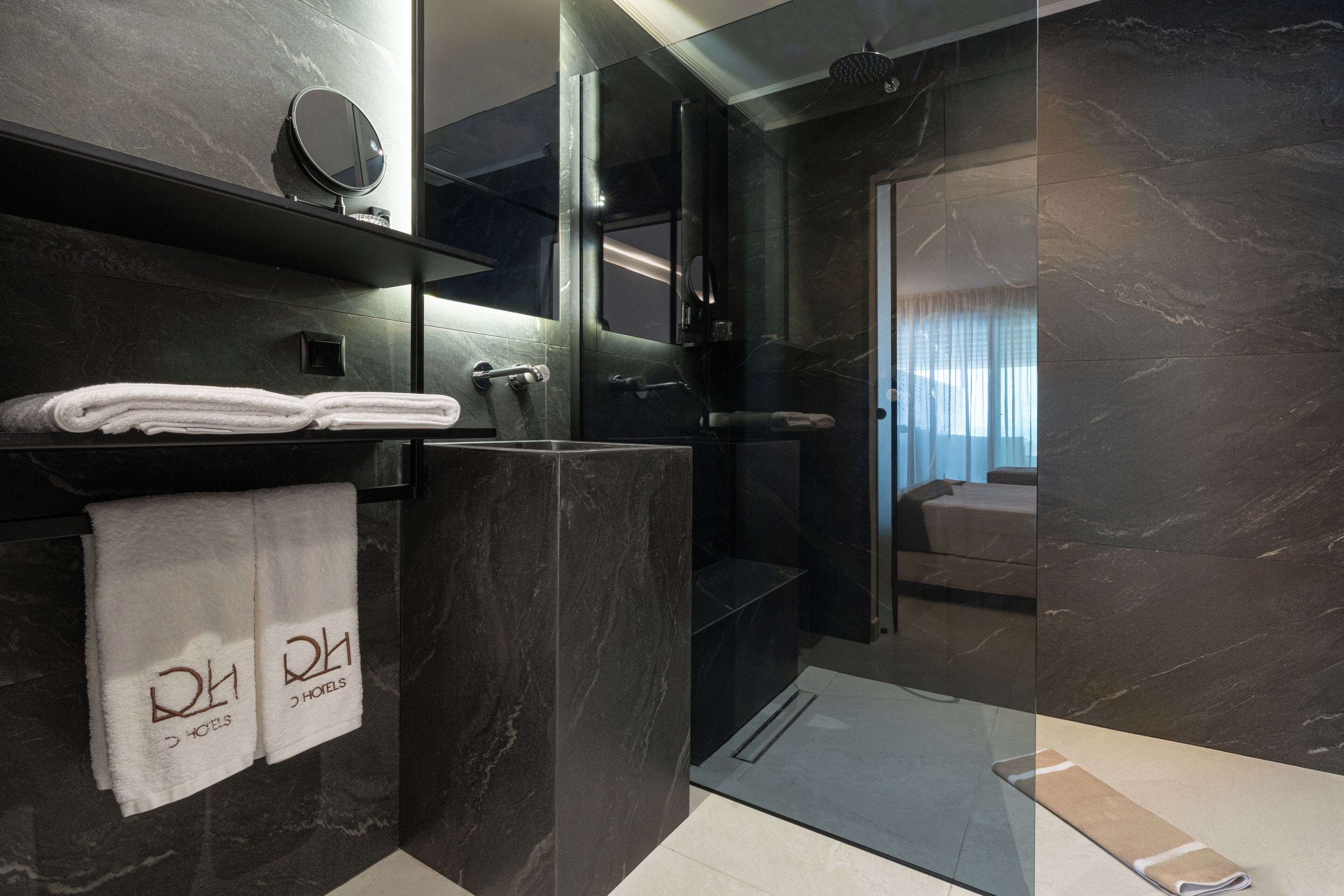A modern bathroom with dark marble walls, a glass shower enclosure, and a visible bedroom with a bed and curtains.