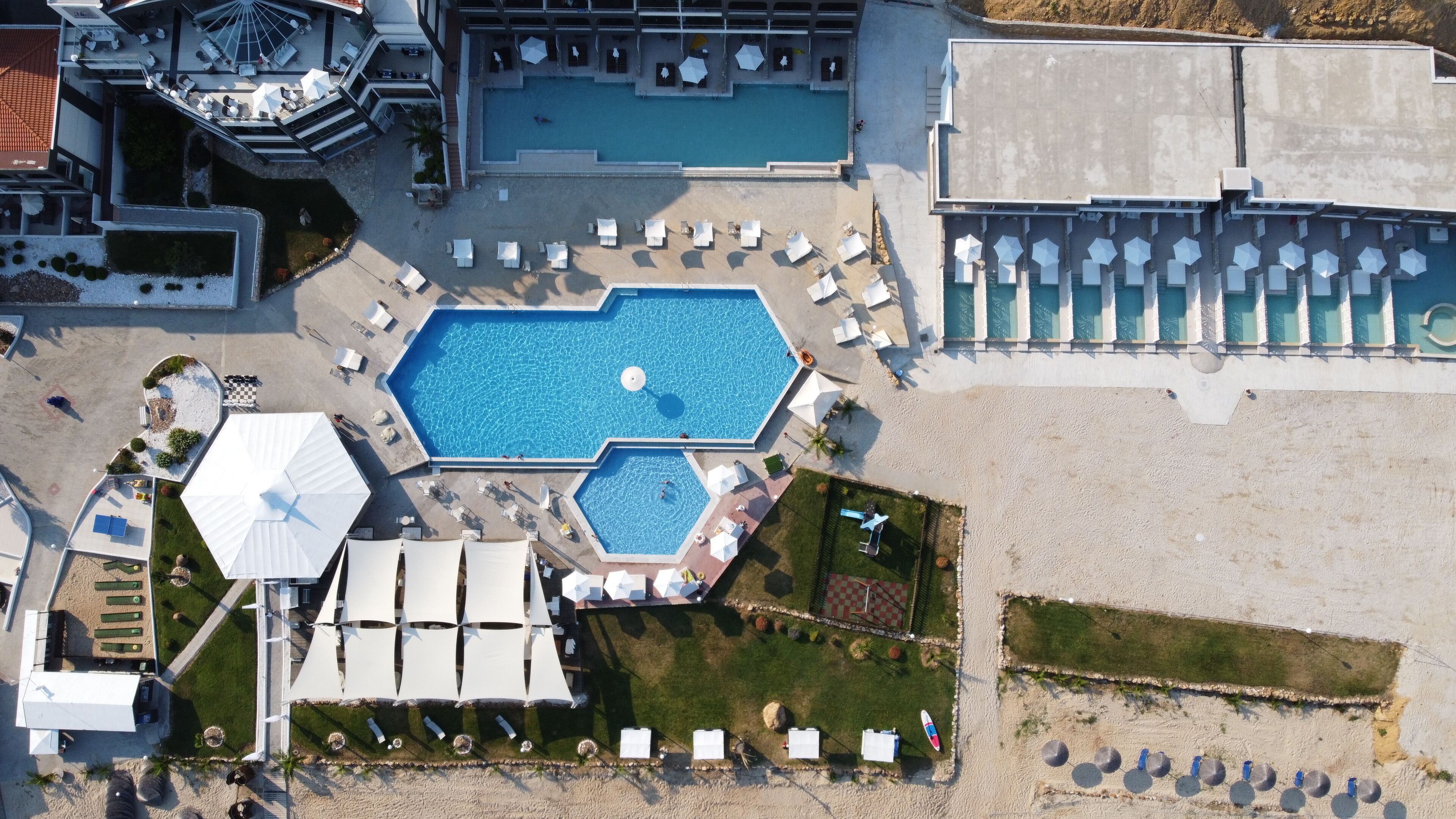 Vista aérea de un hotel con una amplia piscina central, múltiples tumbonas y un jardín bien cuidado.