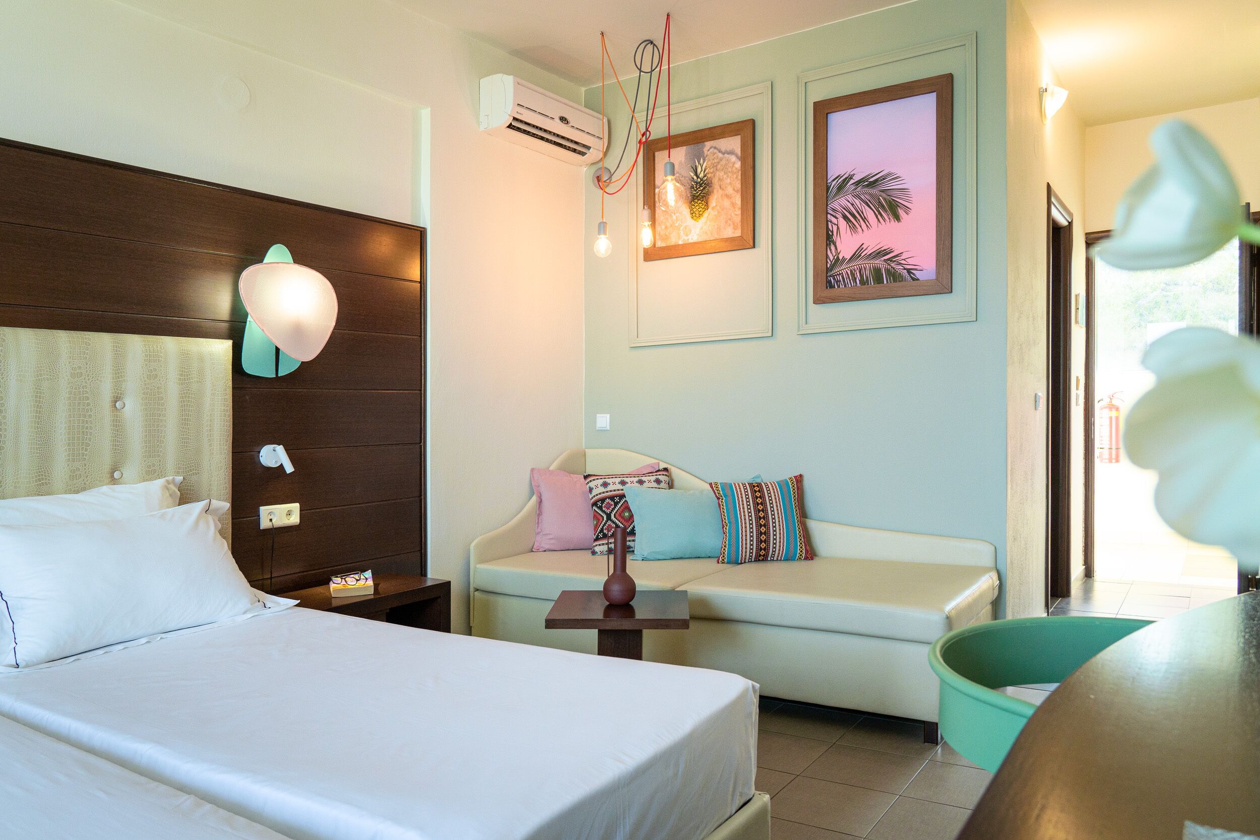A hotel room with a bed, a sofa, a small table, and an air conditioning unit.
