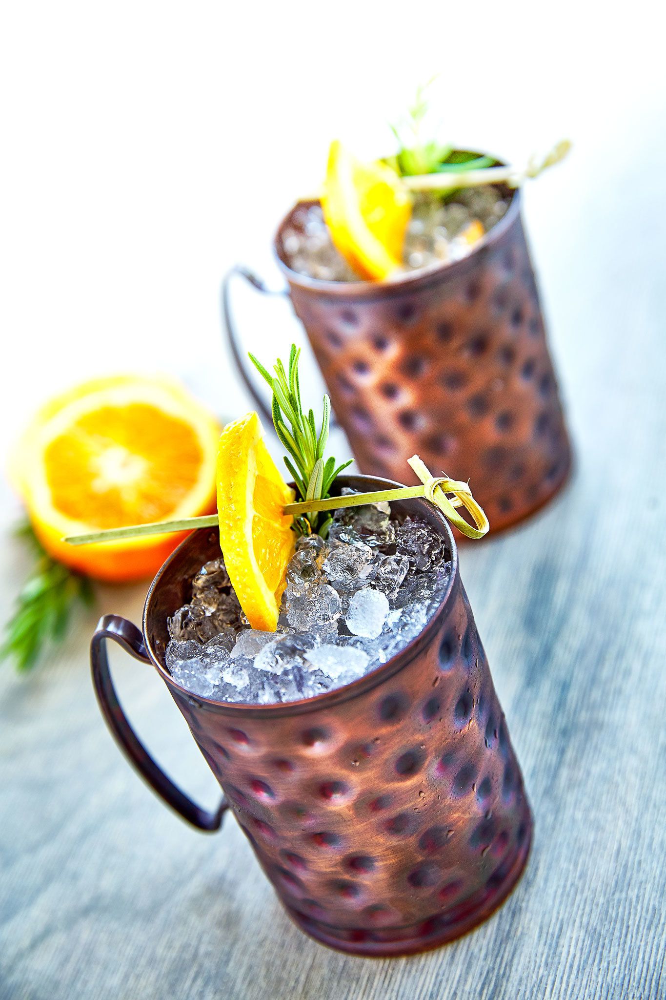 Two copper mugs with ice and garnishes on a wooden surface.