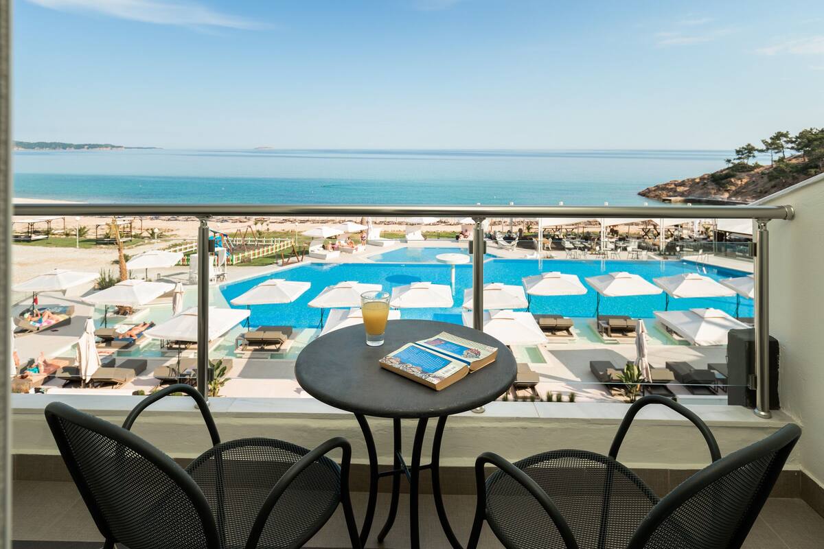 Varanda com vista para uma piscina e praia. Há cadeiras de praia e guarda-sóis.