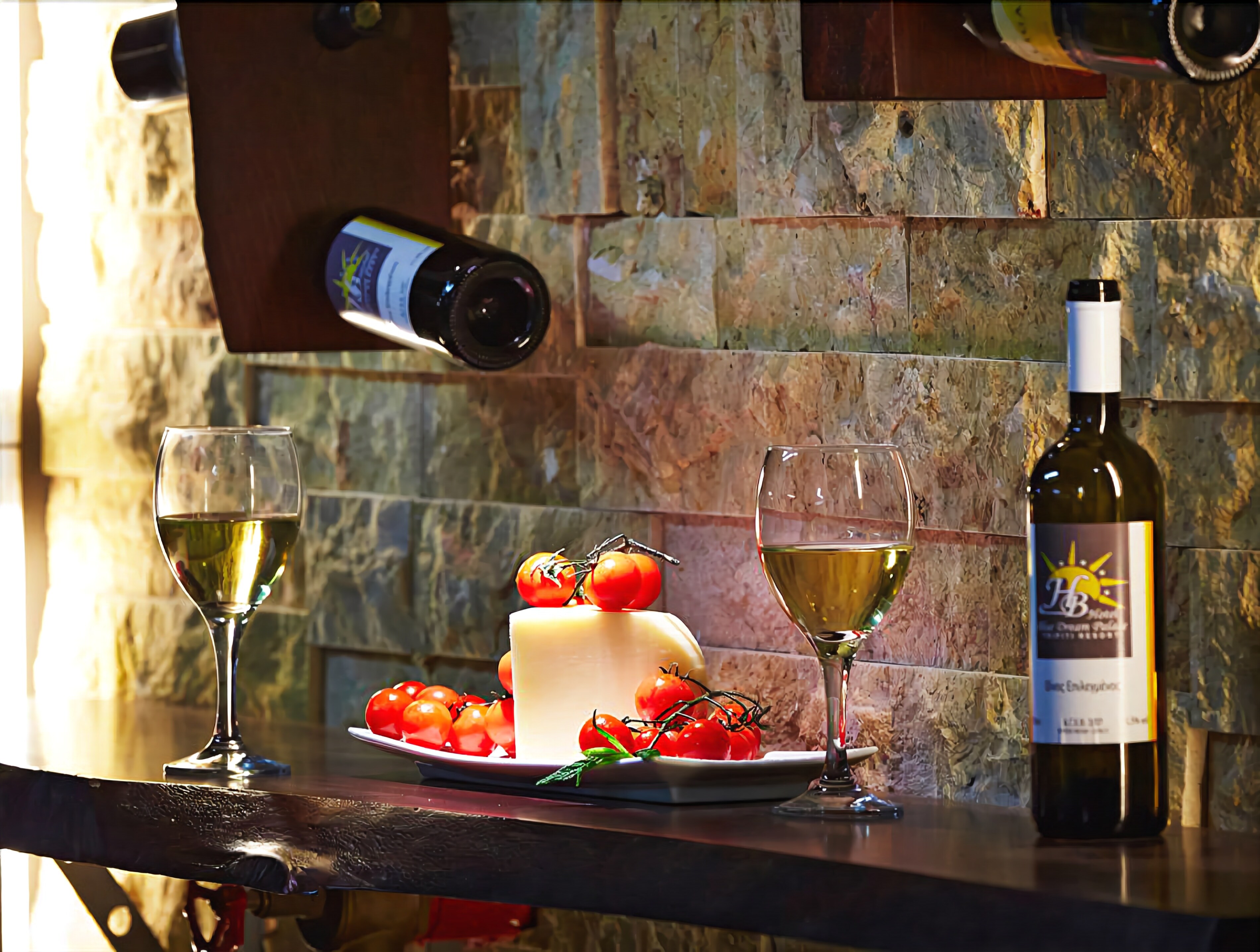 Une bouteille de vin, deux verres de vin blanc, une assiette avec du fromage et des tomates, et une bouteille de vin posés sur une table en bois, le tout adossé à un mur de pierre.