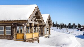 Uma cabana de tronco com varanda de madeira e várias janelas, rodeada por uma paisagem e árvores cobertas de neve.