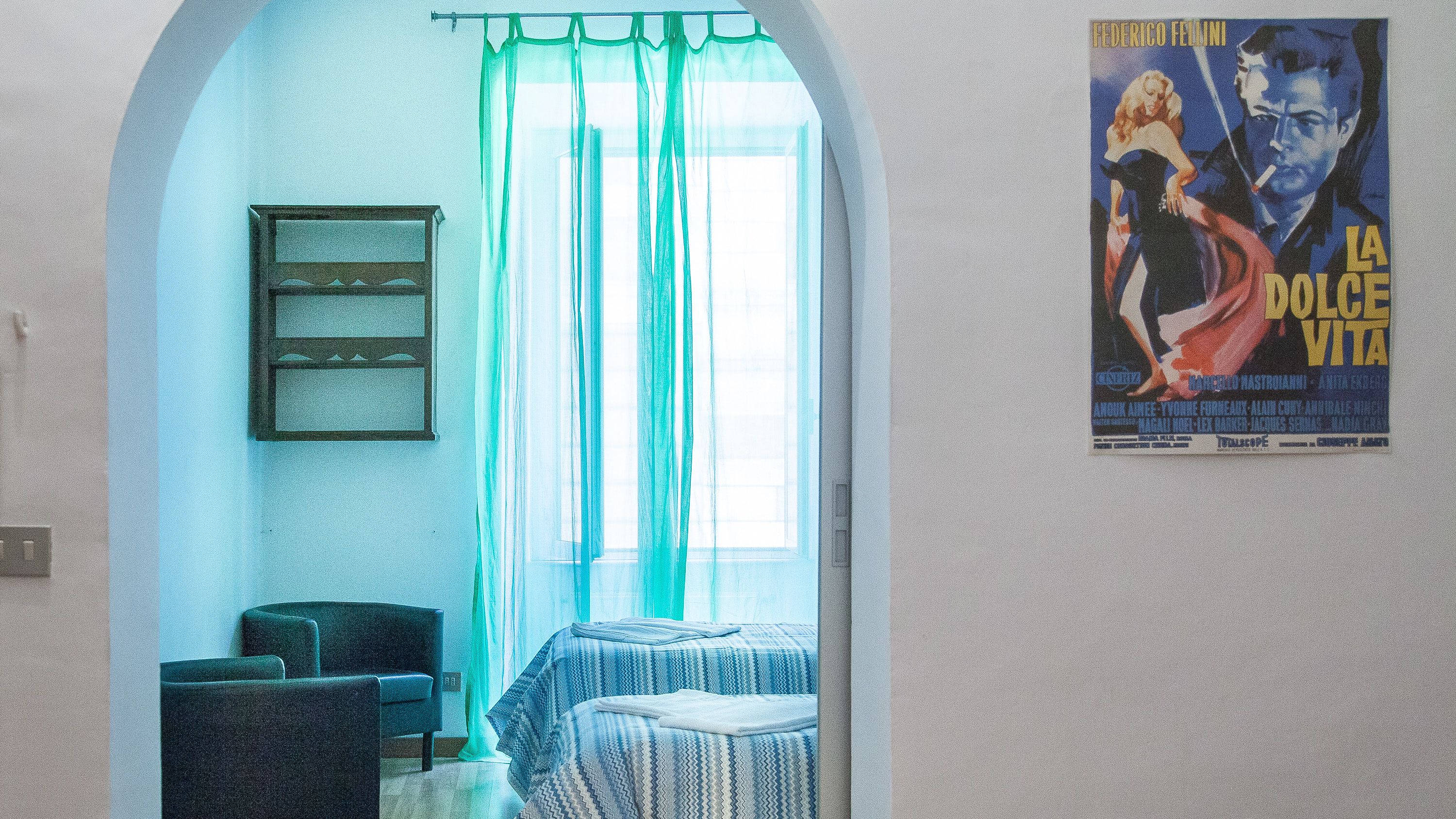 Una habitación con cama, una silla, una ventana con cortinas traslúcidas y un cartel en la pared.
