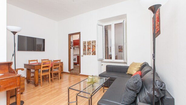 Una sala de estar con una mesa de comedor de madera, sillas, una mesa de centro de vidrio, un sofá negro, una lámpara de pie y un televisor en la pared.