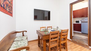 Un comedor con una mesa y sillas de madera, un televisor montado en la pared y una cocina a la vista a través de una puerta abierta.