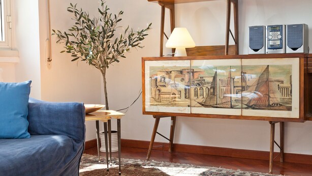 A living room with a blue sofa, a small table with a lamp, a framed painting, and a potted plant. - Rental In Rome Pateras Flower (Rome)