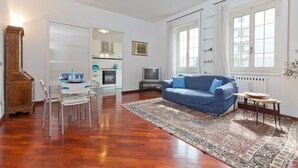 A living room with a blue sofa, a dining table with chairs, a kitchen area, and a large window with curtains. - Rental In Rome Pateras Flower (Rome)