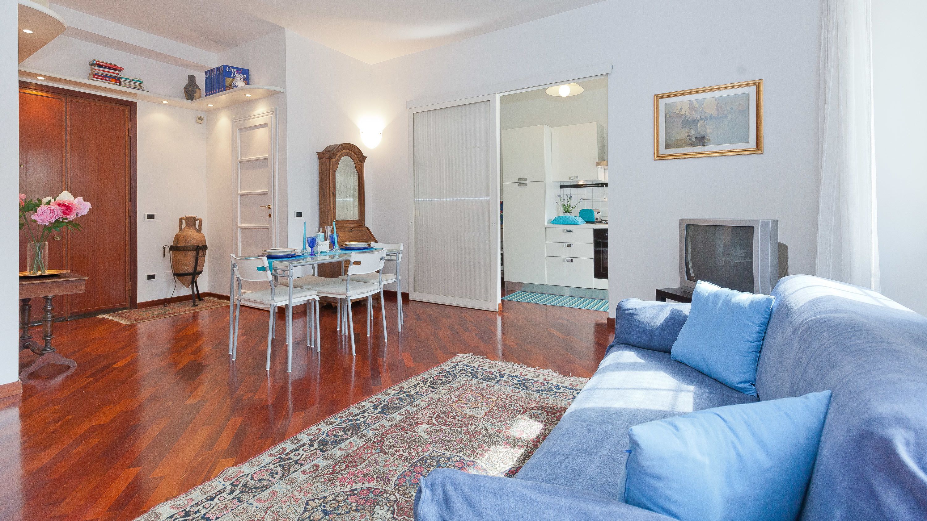 A living room with a sofa, a dining table with chairs, a TV, and a kitchen area in the background.
