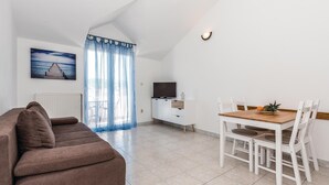 En moderne stue med en brun sofa, et spisebord med hvide stole, et fjernsyn på et hvidt skab, og havudsigt gennem lette gardiner.
