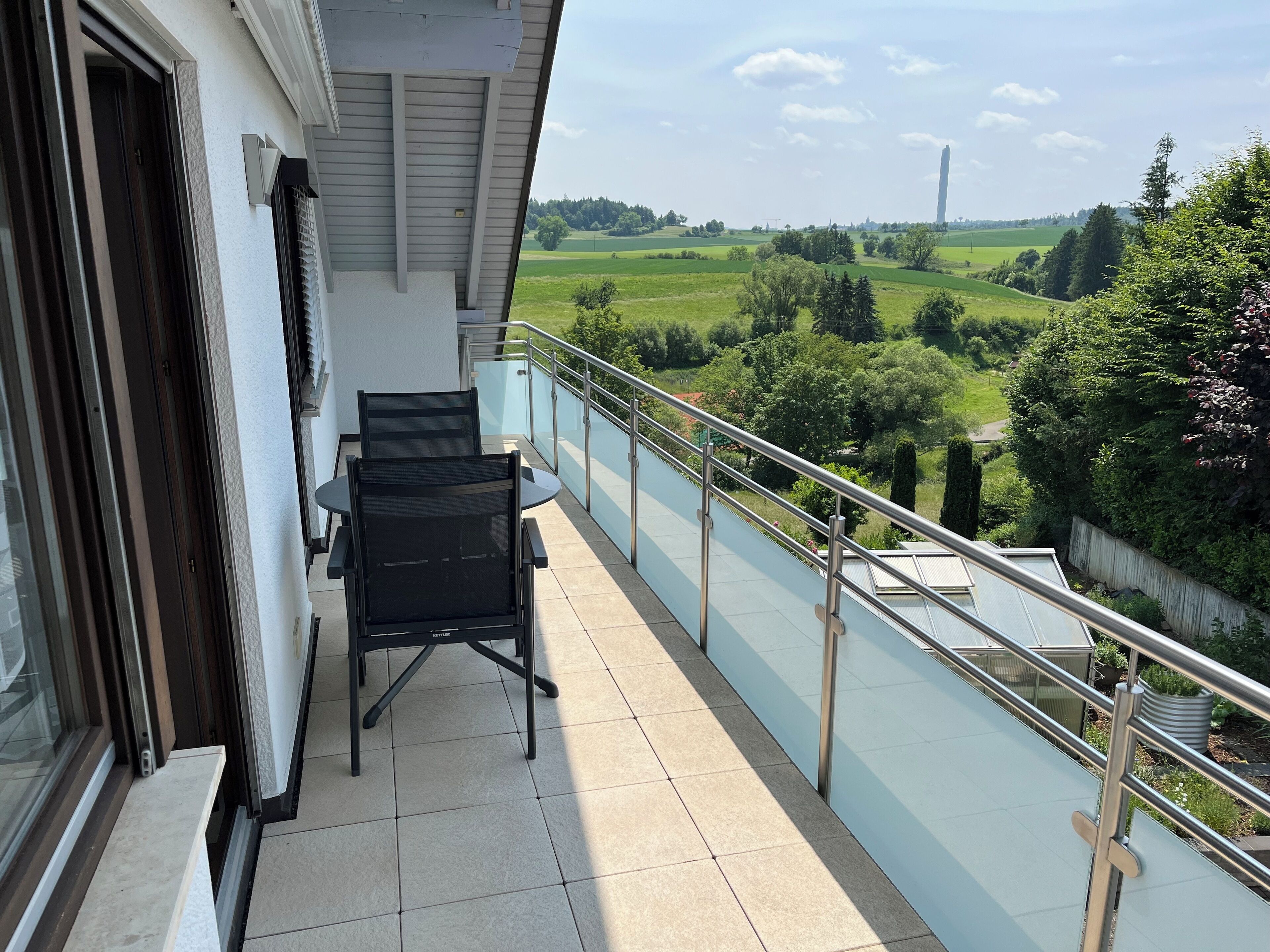 A balcony with a table and chairs, offering a view of a green landscape with trees and a distant tower.