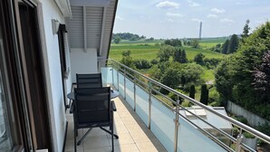 A balcony with a table and chairs, offering a view of a green landscape with trees and a distant tower.