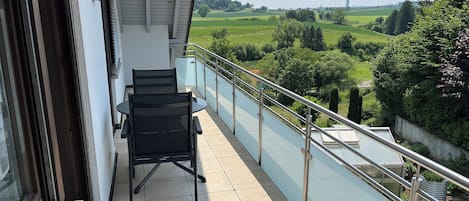 A balcony with a table and chairs, offering a view of a green landscape with trees and a distant tower.