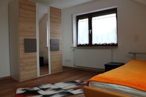 A bedroom with a bed, a window with curtains, a radiator, a wooden wardrobe, and a mirror.